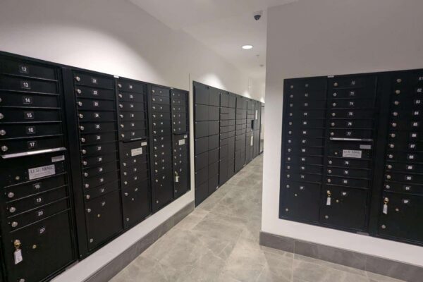 Row of black mailbox lockers with numbered compartments lining a hallway in a modern building corridor, each with a small key lock.