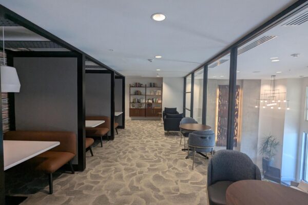 Modern office lounge with booth seating along left wall, round tables on the right, and a glass partition across the room.
