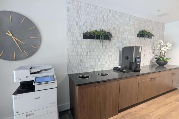 Break room with a large gray wall clock, coffee machine, and hex-tile backsplash above wooden cabinets.