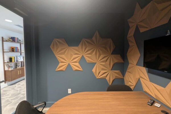 Conference room corner with a dark blue wall featuring brown geometric acoustic panels, an oval wooden table, and a black chair; a mounted TV is on the right wall. A doorway reveals a shelving unit in an adjoining room.