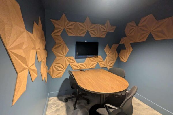 Small meeting room with blue walls and tan geometric acoustic panels, an oval wooden table, and rolling chairs around it.