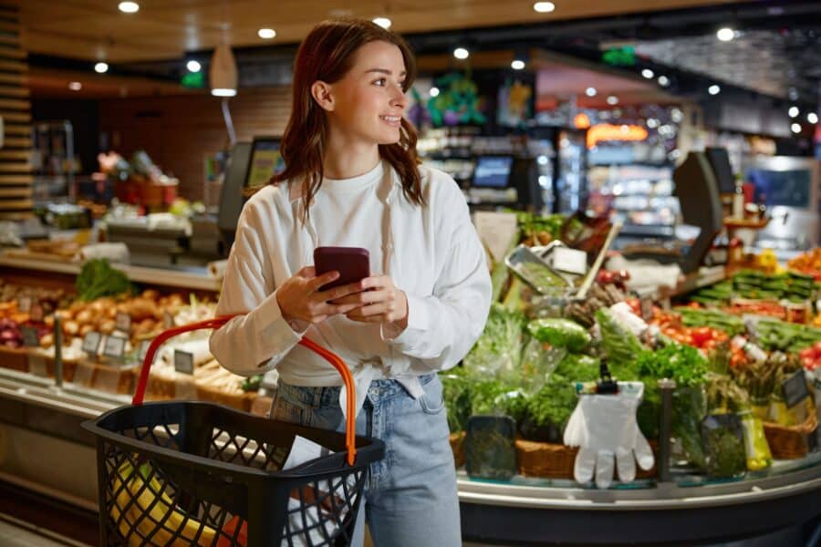 Woman smiling while holding a smartphone in a grocery store, with fresh produce and shelves in the background.